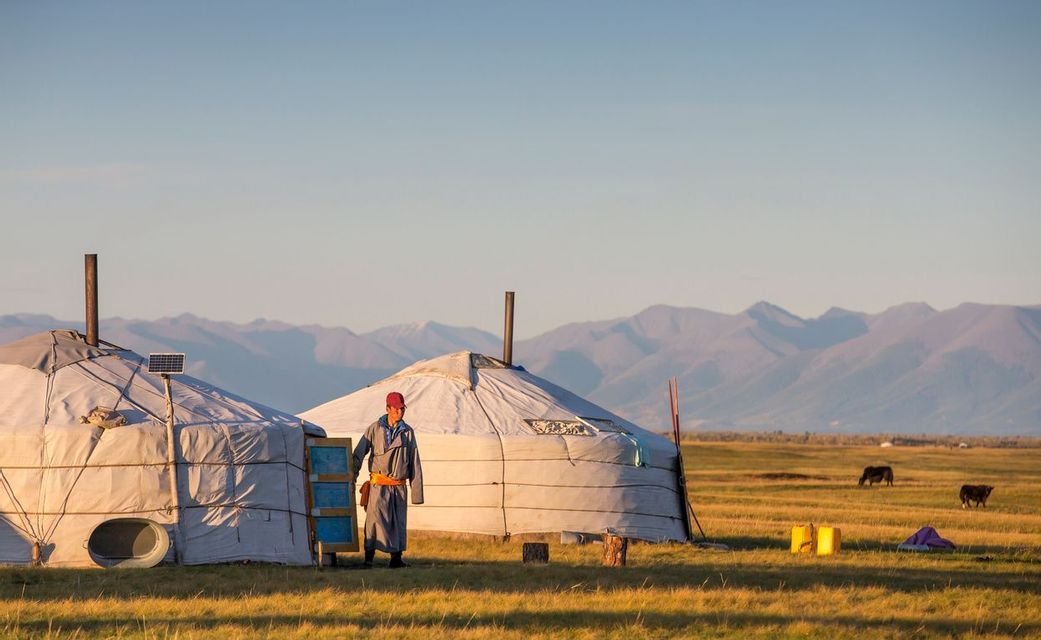 Una persona con ropa tradicional se encuentra junto a dos yurtas en un campo de hierba con una cordillera al fondo.