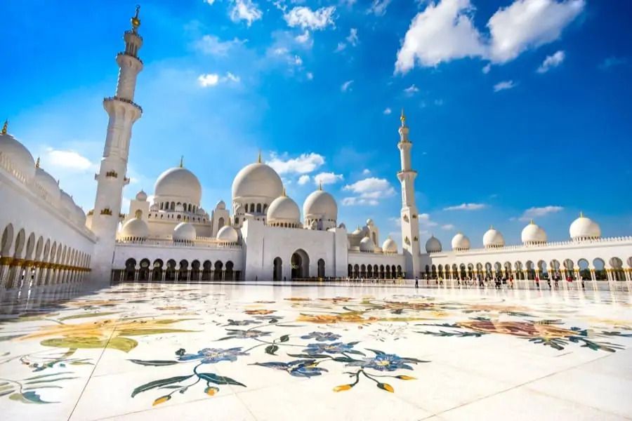 Eine große, weiße Moschee mit mehreren Kuppeln und Minaretten, vom Innenhof aus gesehen, der unter blauem Himmel aufwendige florale Marmoreinlegearbeiten präsentiert.