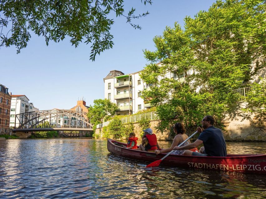 Eine vierköpfige Familie paddelt in einem roten Kanu auf einem Stadtfluss, vorbei an Bäumen und Gebäuden.