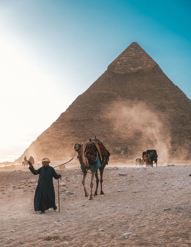 Una persona in abiti tradizionali conduce un cammello attraverso il deserto, con una grande piramide che si staglia sullo sfondo polveroso.