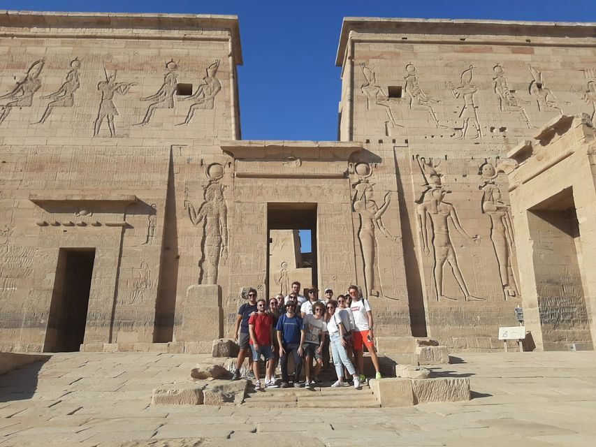 A WeRoad group trip poses for a photo on the steps of a large ancient Egyptian temple with hieroglyphic carvings under a clear blue sky.
