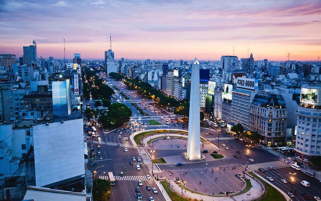 Una vista aerea di una città al tramonto, con un grande obelisco in una piazza circondata dal traffico su un ampio viale.