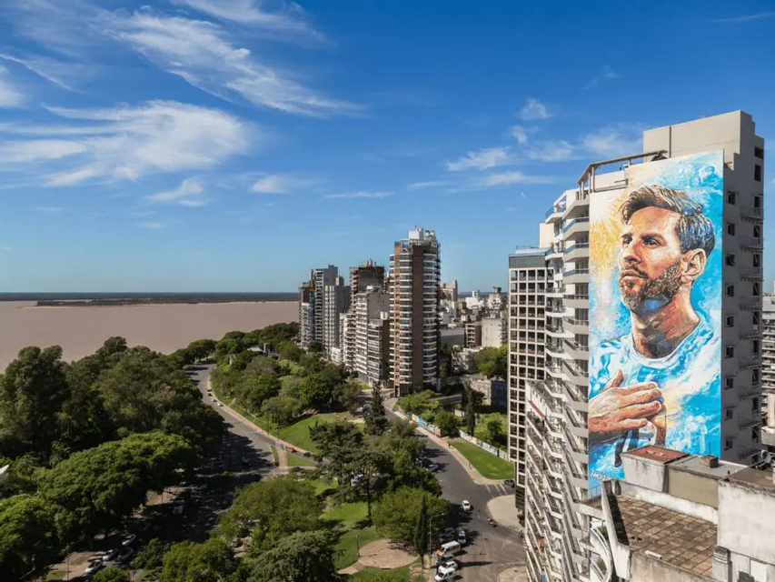 Un grande murale di Lionel Messi su un grattacielo, con vista su un paesaggio urbano verde e un ampio fiume sotto un cielo blu.