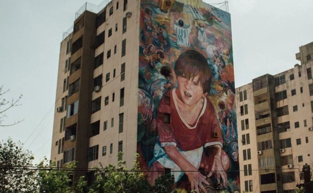 Un enorme murale colorato di un giovane ragazzo con una maglietta rossa è dipinto sul fianco di un condominio a più piani.