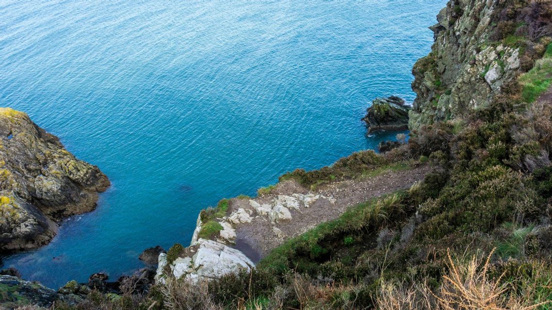 Ein schmaler Feldweg führt entlang einer rauen, grasbewachsenen Klippe mit Blick auf das blaue Meer.