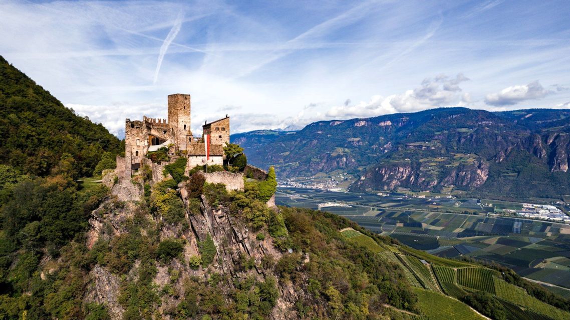 Eine mittelalterliche Steinburg thront auf einer felsigen, baumbestandenen Klippe über einem weiten Tal mit Feldern und Bergen unter blauem Himmel.