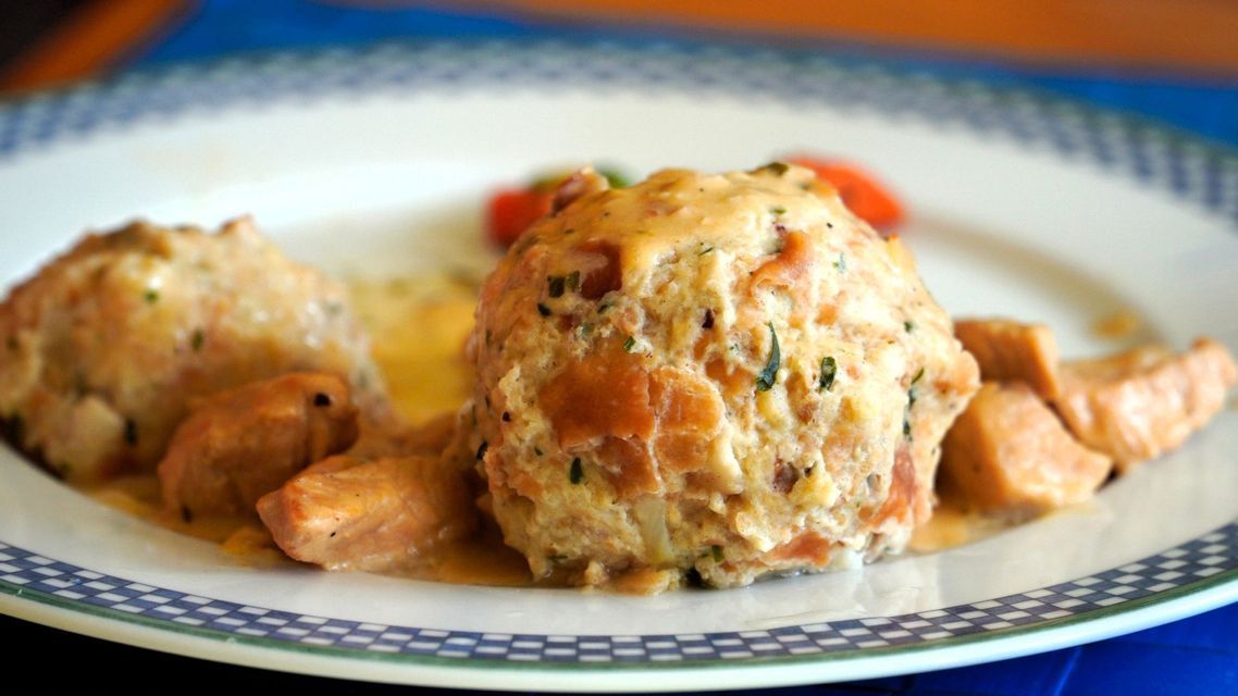 Nahaufnahme von zwei großen Semmelknödeln und Fleischstücken, bedeckt mit einer cremigen Soße auf einem dekorativen Teller.