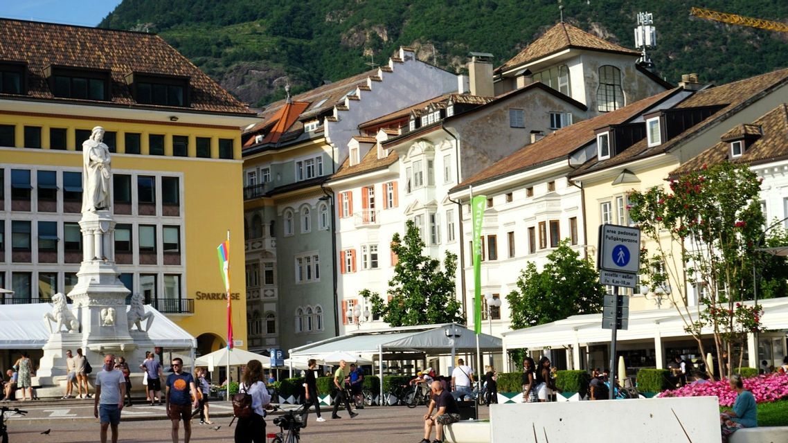 Menschen spazieren durch einen sonnigen europäischen Stadtplatz mit einer markanten weißen Statue, farbenfrohen Gebäuden und einem Berg im Hintergrund.