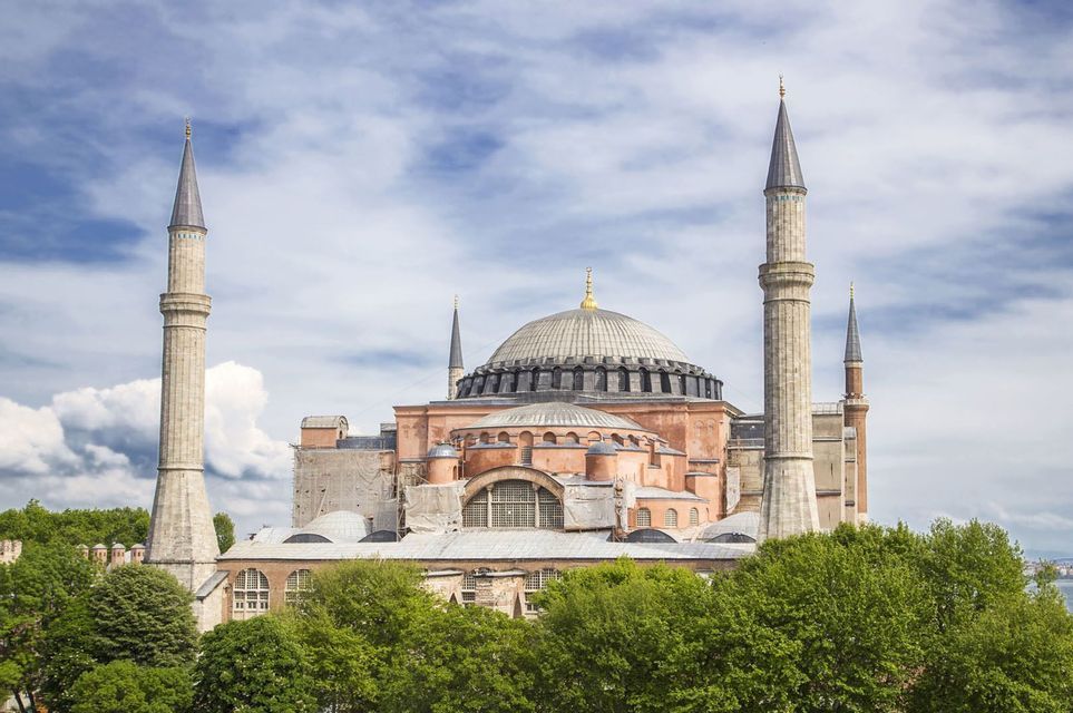 Una grande moschea con una cupola centrale e alti minareti si erge sopra le cime degli alberi lussureggianti sotto un cielo blu con nuvole bianche.