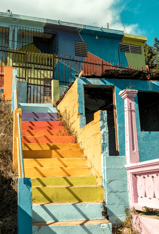 Una escalera exterior pintada con un degradado de arcoíris sube a un edificio colorido bajo un cielo parcialmente nublado.