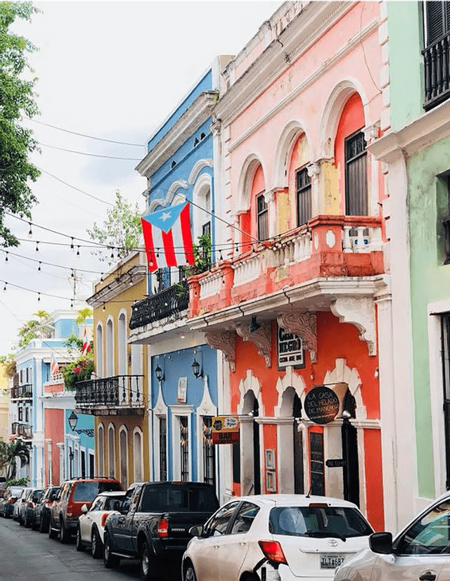 Una bandera puertorriqueña cuelga de un balcón en una calle llena de edificios coloniales coloridos y coches aparcados.