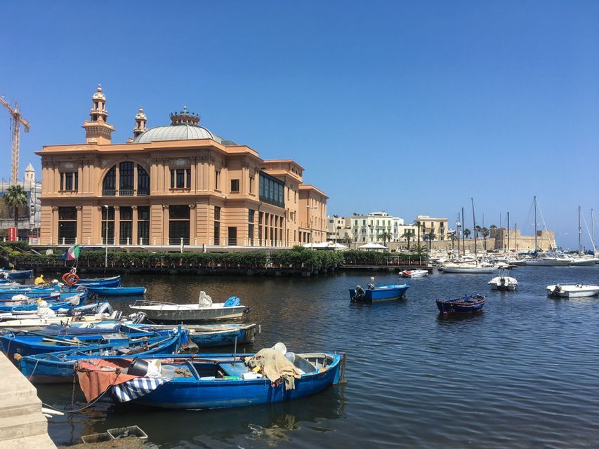 Un porto con piccole barche blu ormeggiate di fronte a un grande edificio color pesca riccamente decorato, sotto un cielo azzurro e sereno.