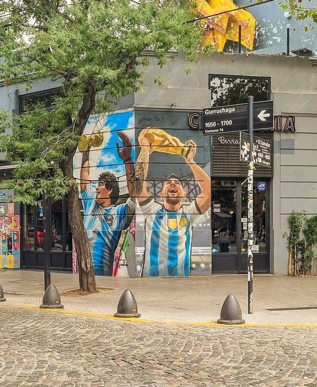 Un grande murale all'angolo di un edificio raffigura i calciatori Maradona e Messi che festeggiano con i trofei della Coppa del Mondo.