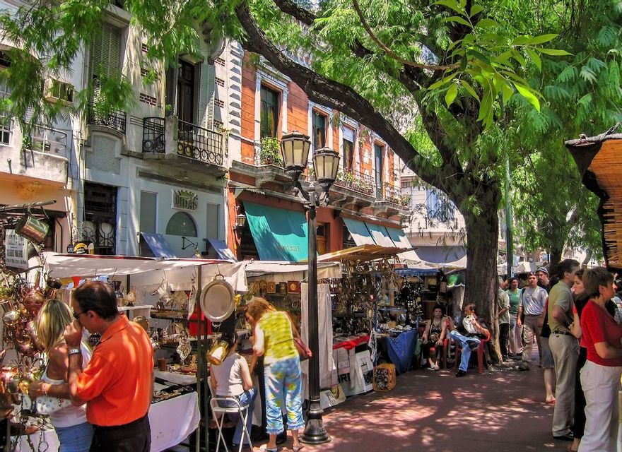 Un vivace mercato all'aperto in una giornata di sole, con persone che curiosano tra le bancarelle sotto alberi frondosi accanto a edifici storici.