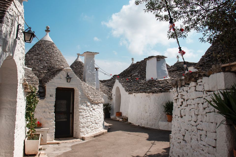 Casas de piedra encaladas con techos cónicos bordean un callejón estrecho y pavimentado bajo un cielo azul con algunas nubes.