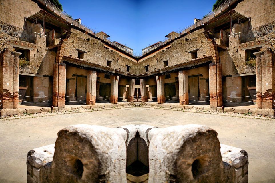 Una vista specchiata e simmetrica del cortile di un'antica rovina con pilastri in mattoni e muri in pietra sotto un cielo azzurro chiaro.