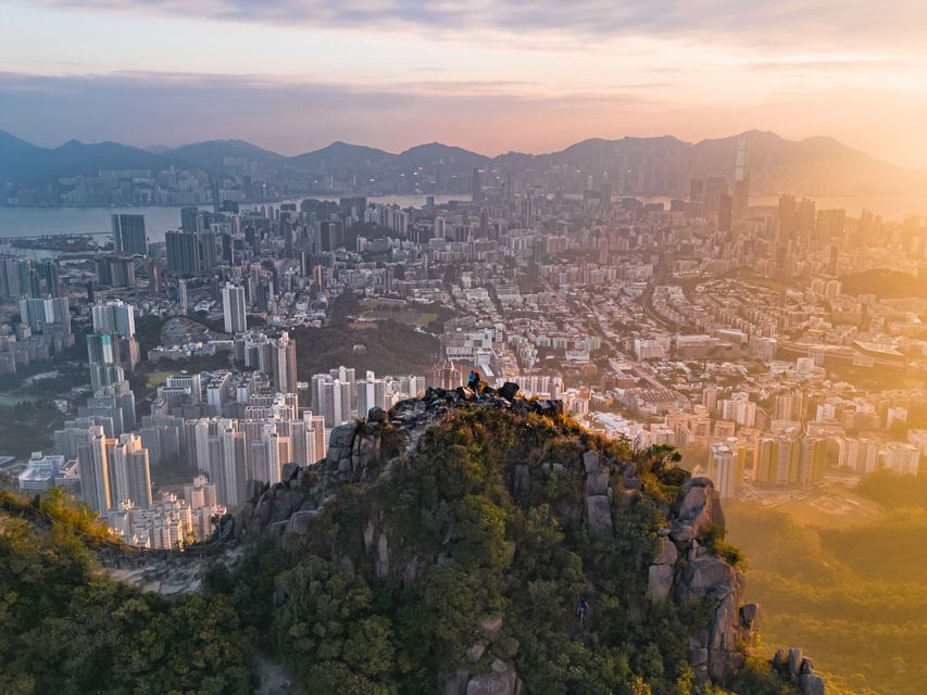 Two people on a rocky mountain peak overlooking a sprawling cityscape at sunrise, with hazy mountains in the background.