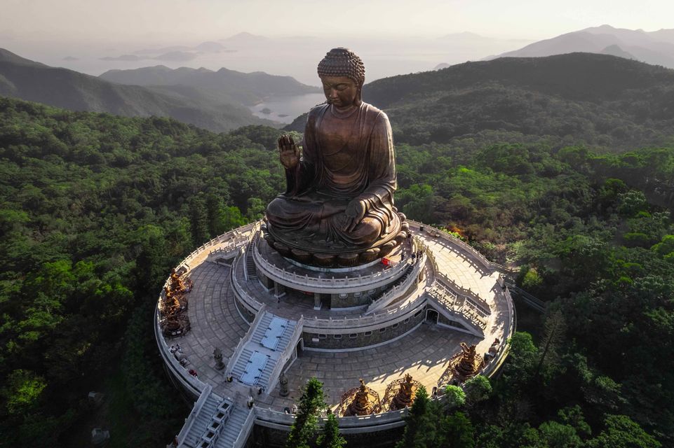 An aerial view of a large bronze Buddha statue seated on a tiered platform, surrounded by lush green mountains and distant water.