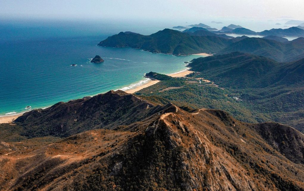 An aerial view from a brown mountain ridge overlooking a coastline with a sandy beach, green hills, and turquoise water under a hazy sky.