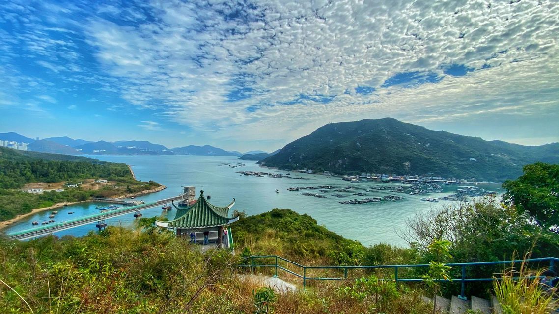 A scenic overlook with a green-roofed pavilion, viewing a bay filled with fish farms and surrounded by lush green mountains under a cloudy sky.