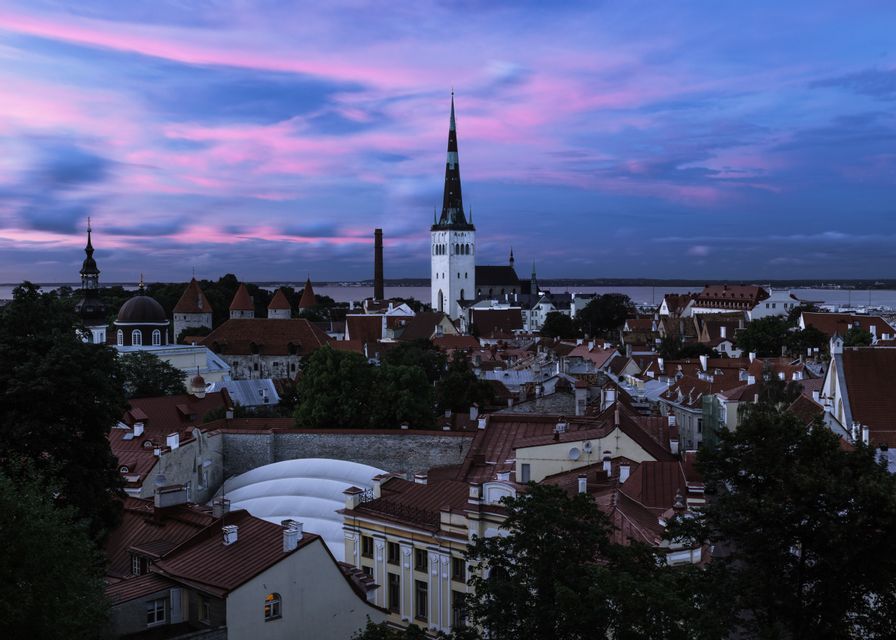 Un horizonte de ciudad europea con un prominente campanario de iglesia y edificios de tejados rojos bajo un cielo de atardecer rosa y morado.
