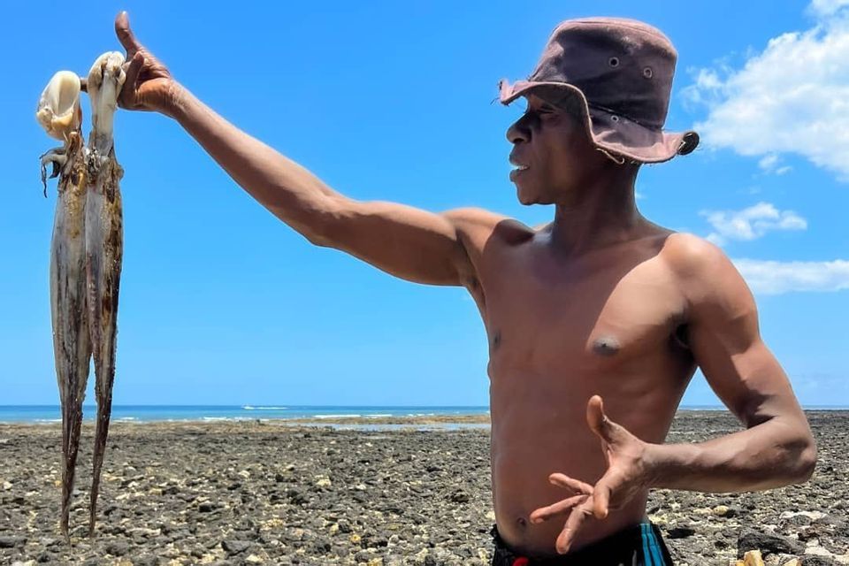 Un uomo con un cappello a secchiello mostra un polpo appena pescato su una spiaggia rocciosa sotto un cielo azzurro e limpido.