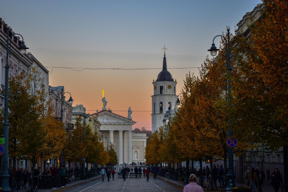 Una strada ampia e alberata con foliage autunnale conduce a una cattedrale e un campanile al tramonto, con persone che camminano in lontananza.