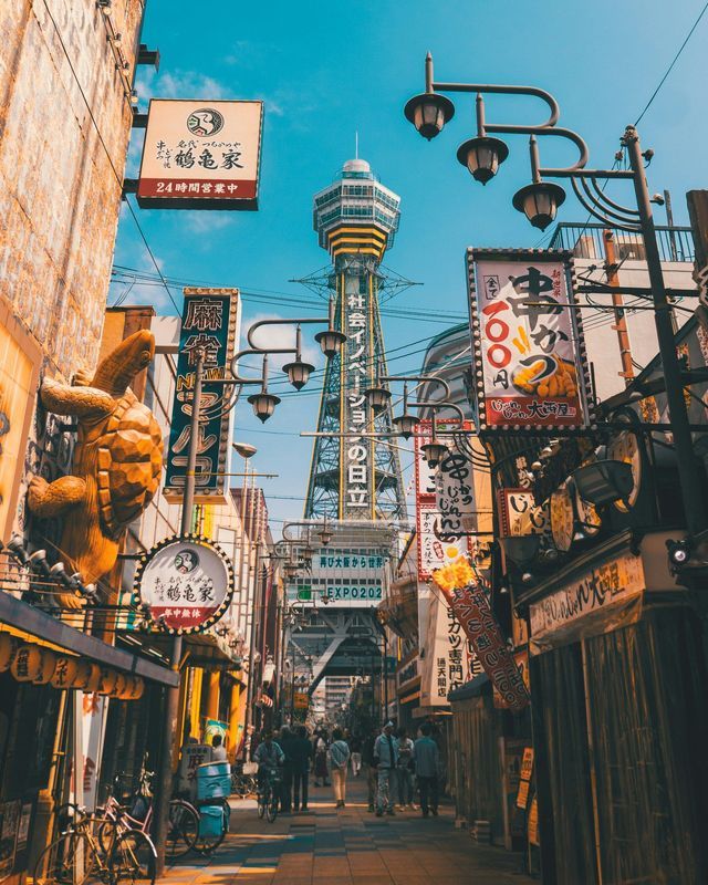 A view down a narrow city street crowded with Japanese signs, with a tall observation tower in the distance under a blue sky.