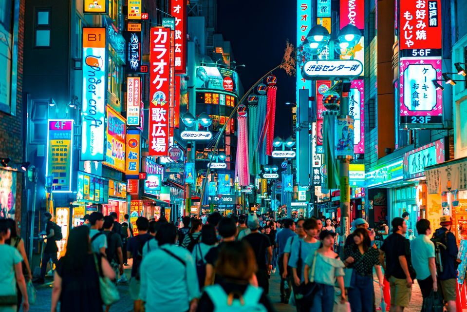 A large crowd of people walks down a narrow city street at night, illuminated by many colorful neon signs.