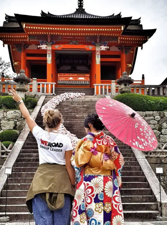 A WeRoad group leader makes a peace sign next to a woman in a kimono at the base of stairs leading to a Japanese temple.