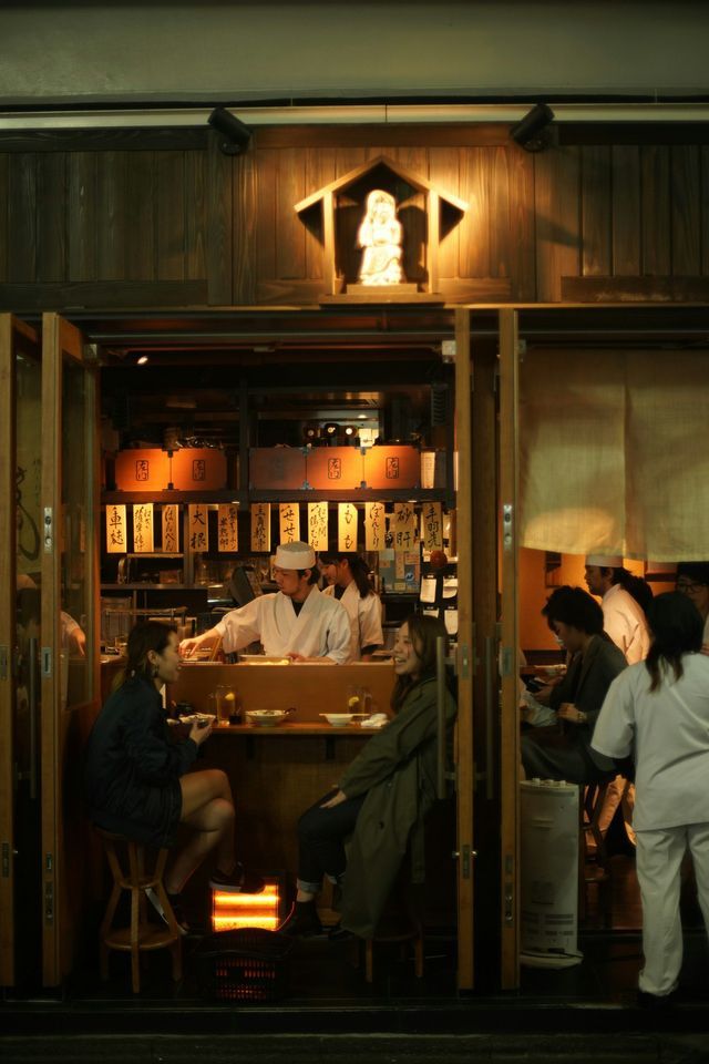 Two women on a WeRoad group trip dining at a traditional Japanese restaurant at night, viewed from the outside.