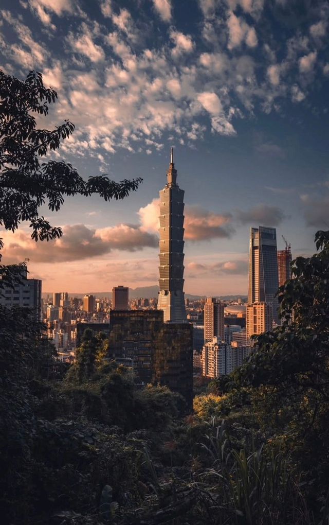 Un alto grattacielo a gradoni si staglia sullo skyline di una città al tramonto, visto da una foresta scura e rigogliosa.