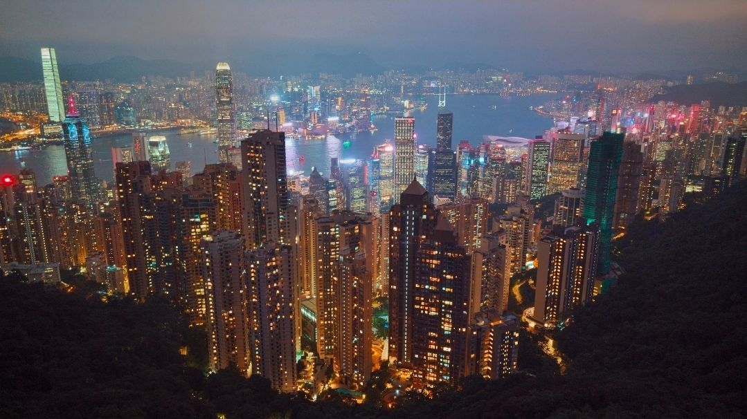 Una veduta aerea di un paesaggio urbano denso con grattacieli illuminati che circondano un porto al tramonto, con montagne sullo sfondo.