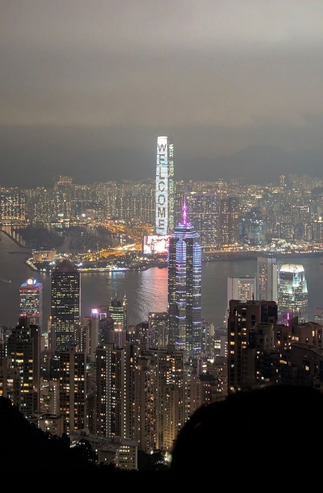 Uno skyline cittadino esteso visto da un punto di osservazione elevato di notte, con un grattacielo che mostra la parola 'BENVENUTI' in luci verticali.