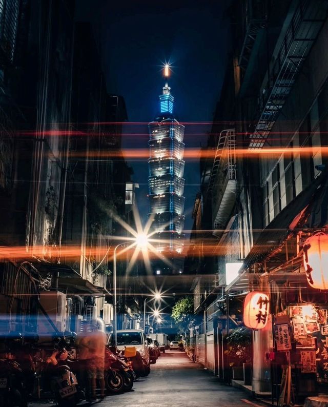 Un grattacielo alto e illuminato visto da uno stretto vicolo cittadino di notte, con scooter parcheggiati, lanterne luminose e scie luminose.