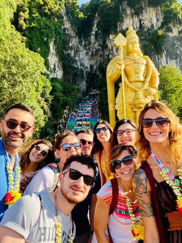 Un viaggio di gruppo WeRoad scatta un selfie davanti a una grande statua dorata e una scala colorata che sale su una scogliera rigogliosa e rocciosa.