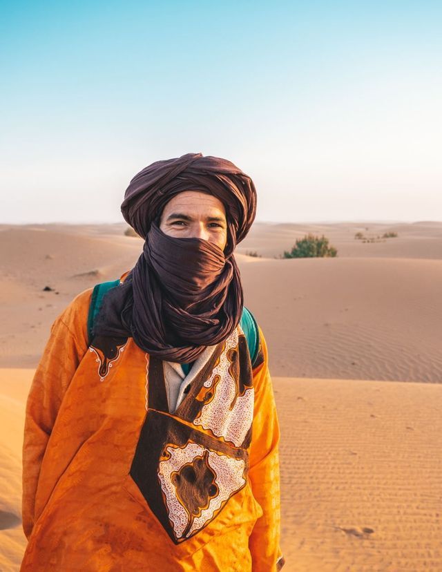 Un uomo che indossa un turbante marrone che gli copre parzialmente il volto e una tunica a motivi arancioni si trova nel deserto.