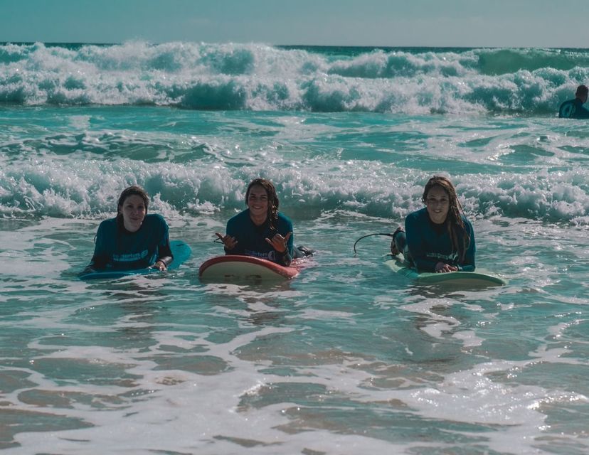 Tre donne del gruppo WeRoad sorridono sdraiate sulle tavole da surf in mare, con le onde che si infrangono dietro di loro.