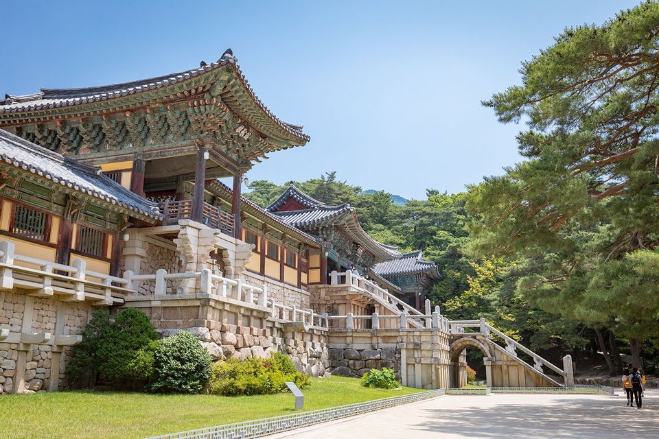 Un tradizionale tempio dell'Asia orientale, con tetti in tegole riccamente decorate, si erge su una fondazione in pietra con una scalinata, circondato da alberi rigogliosi.