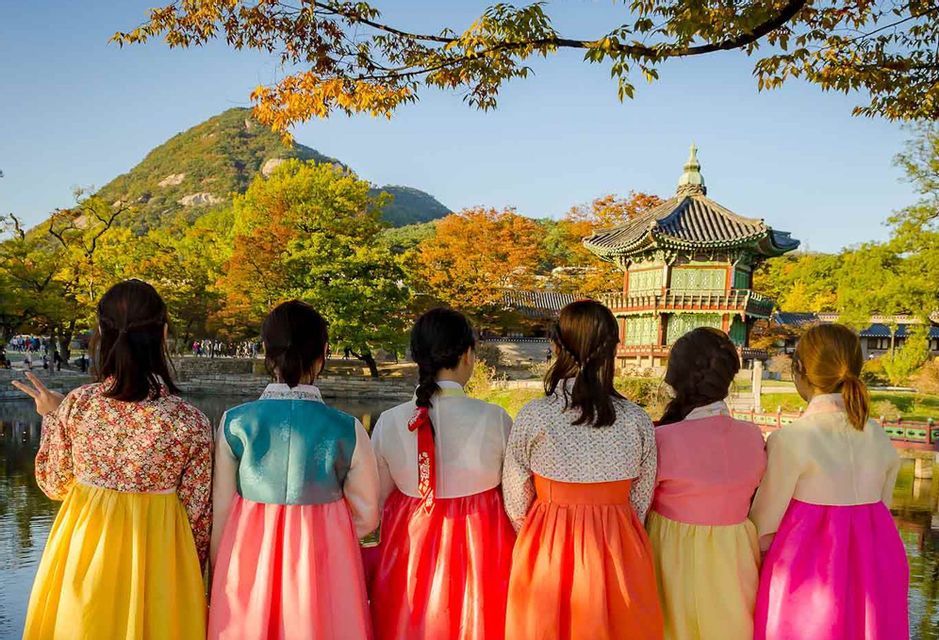 Un viaggio di gruppo WeRoad con donne in colorati abiti tradizionali hanbok che ammirano una pagoda coreana con alberi autunnali sullo sfondo.