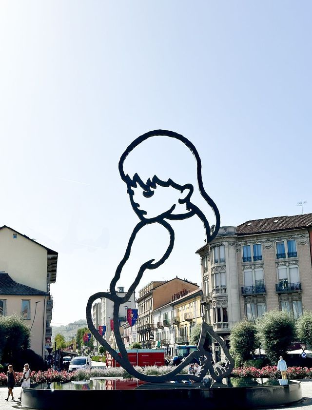 Una grande scultura a sagoma nera di una persona seduta in una fontana in una piazza, con edifici sullo sfondo.