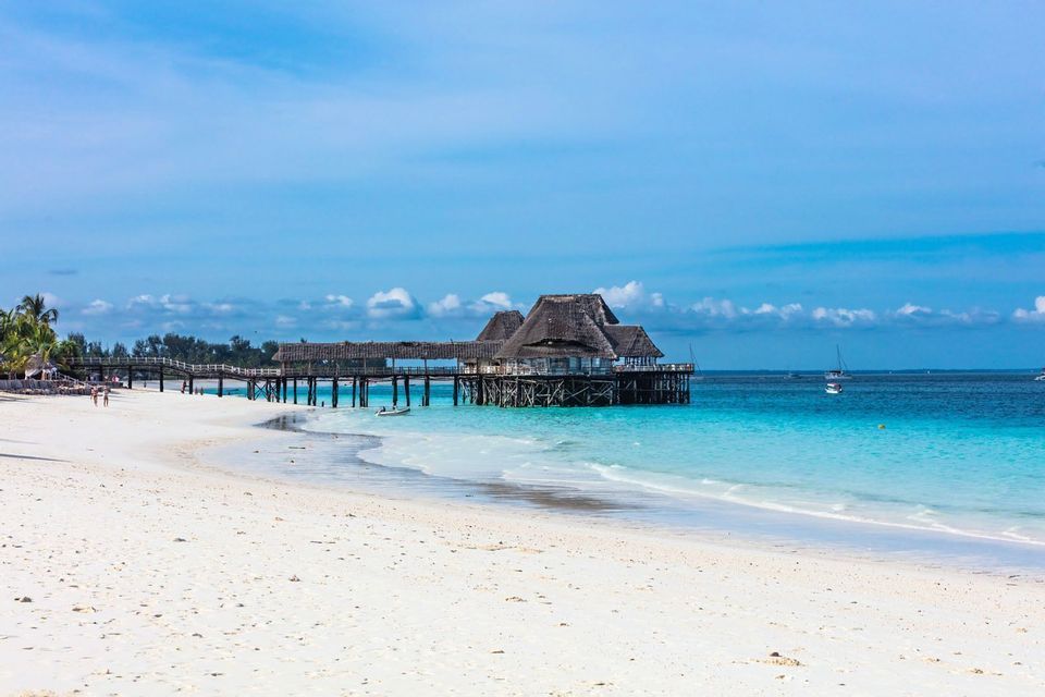 Un bâtiment en bois avec un toit de chaume sur pilotis se dresse au bout d'une jetée au-dessus d'une eau turquoise, à côté d'une plage de sable blanc.