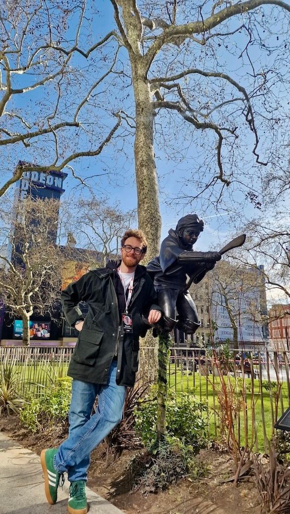 Un uomo sorridente con gli occhiali posa accanto a una statua di bronzo di una figura che vola su una scopa in una piazza della città.