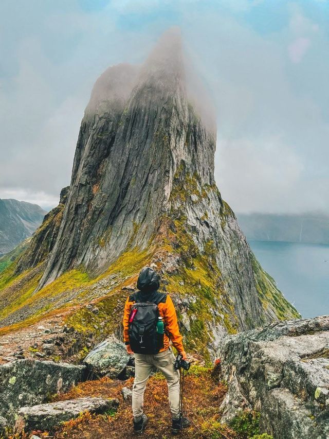A hiker with a backpack and camera stands on a rocky, mossy ridge, looking up at a tall mountain peak shrouded in mist.