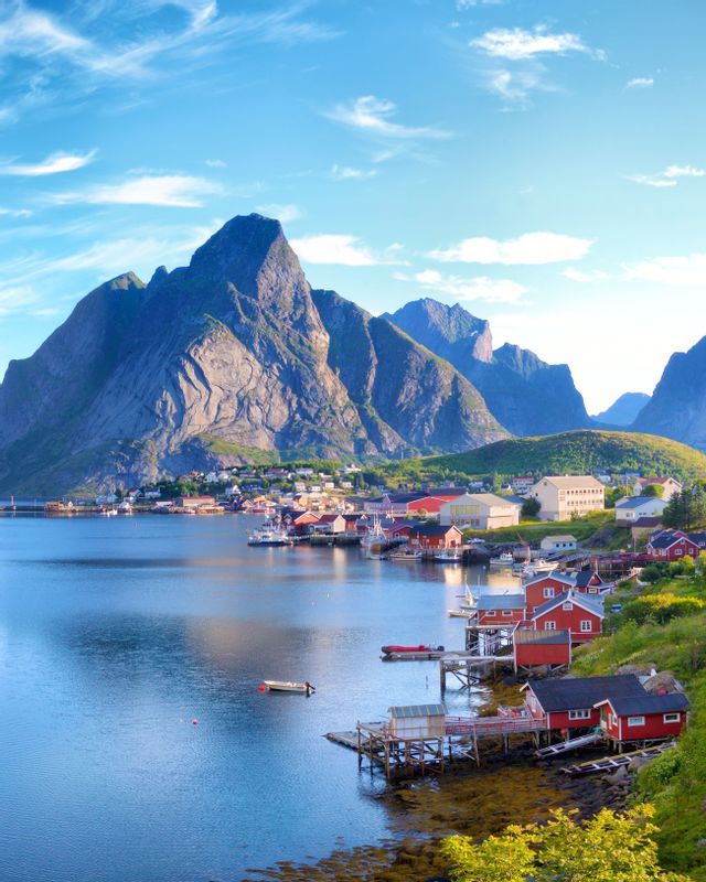 A coastal village with red wooden houses on stilts sits on the edge of calm blue water, with large, steep mountains in the background.