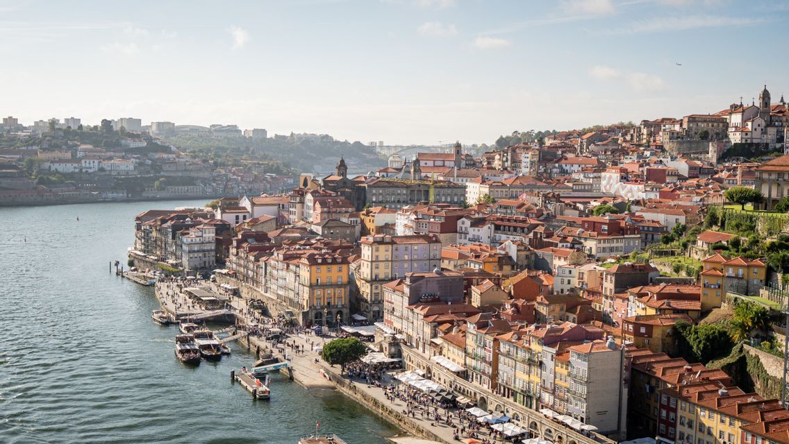 A high-angle view of a city with colorful buildings and terracotta roofs built on a hillside next to a wide river.