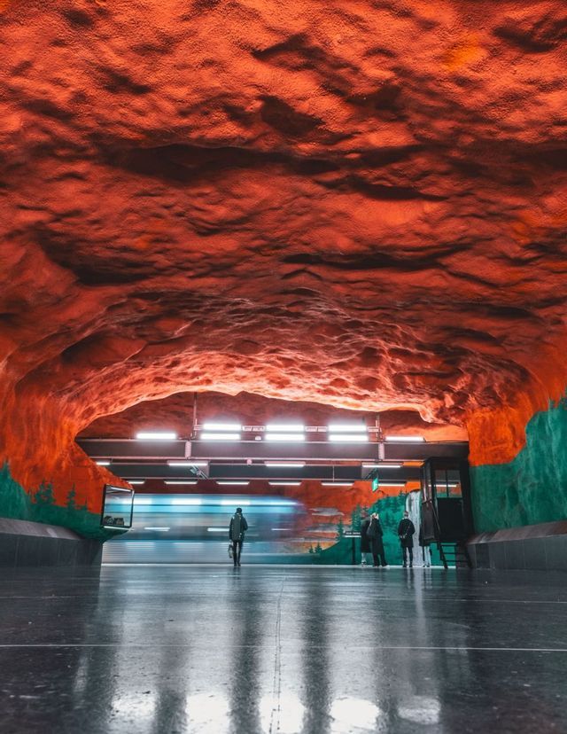 Un treno sfreccia in una stazione della metropolitana con un soffitto rosso che ricorda una caverna e pareti dipinte, mentre i passeggeri attendono sulla banchina riflettente.
