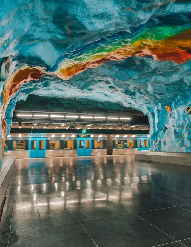 Un train de métro sur le quai d'une station, avec un plafond en forme de grotte peint en bleu et orné d'une bande arc-en-ciel colorée.