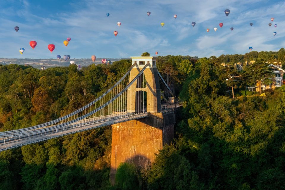 Decine di mongolfiere colorate fluttuano in un cielo blu sopra un grande ponte sospeso circondato da fitti alberi verdi.