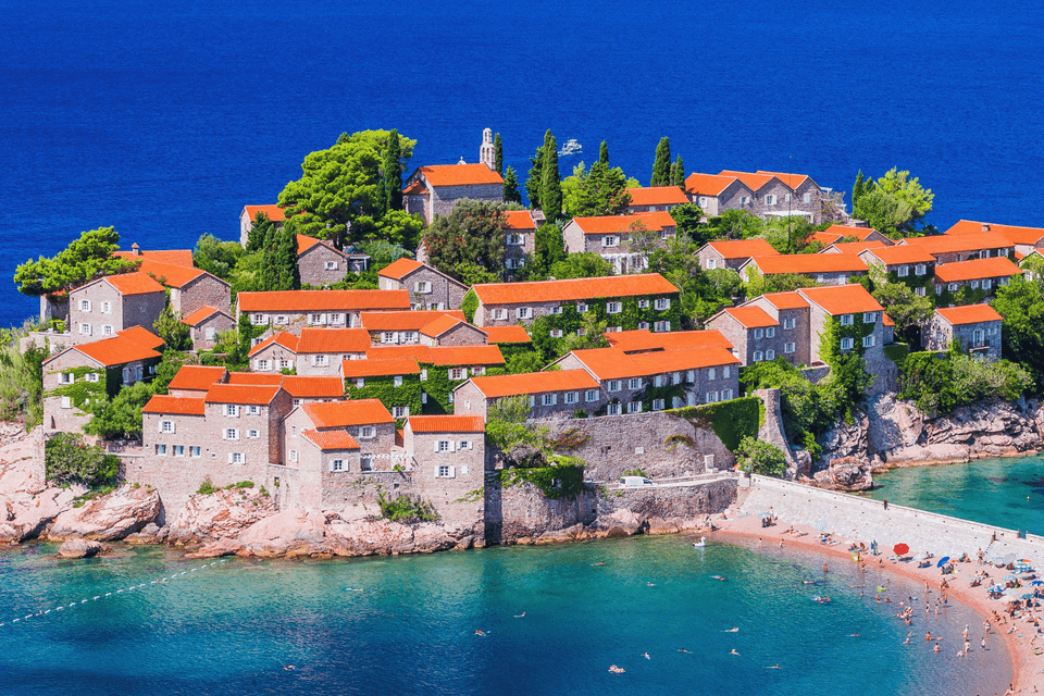 Veduta dall'alto di un villaggio isolano con case in pietra e tetti rossi, delimitato da una spiaggia sabbiosa e dal mare blu.